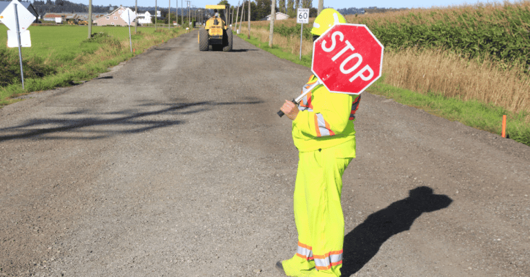 A construction worker holding a stop sign on a dirt road, with loader/tractor on the road up ahead.