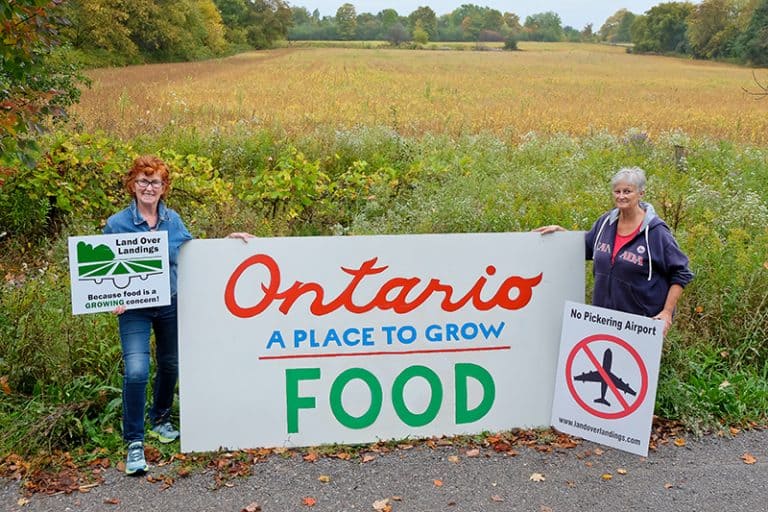 Two women standing next to a sign that says Ontario is a place to grow food on a farm.