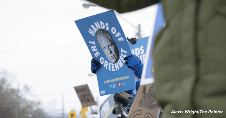 A person holding a protest sign featuring the words "hands off our Greenbelt" and a headshot photo Doug Ford