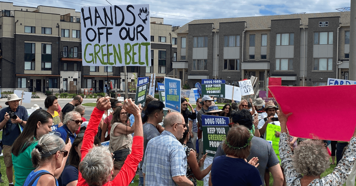 A group of people holding "Hands off our Greenbelt" protest signs in front of an Ontario building.