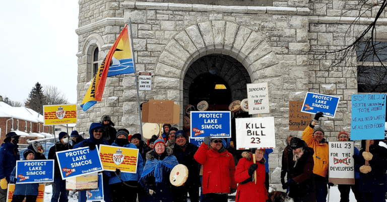 A group of people holding protest signs, "protect Lake Simcoe" and "stop Bill 23", in front of a building.
