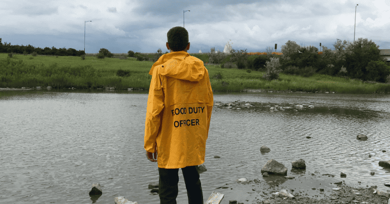 A person in a yellow raincoat that says "food duty officer" on the back, standing near a body of water.