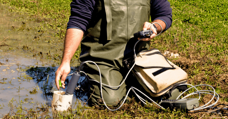 A man kneeling in a puddle testing groundwater with a small, portable machine.