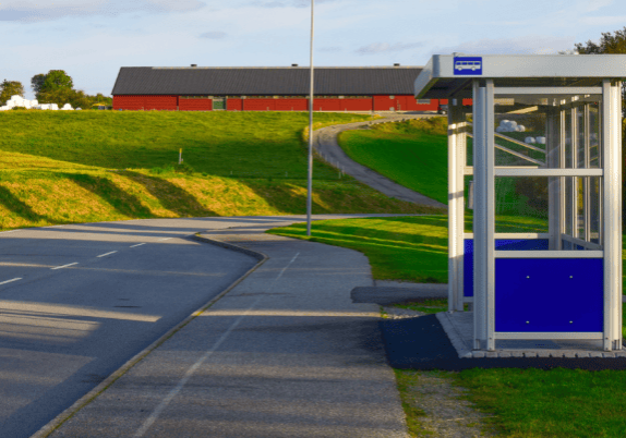 A bus stop on the side of a road with grass and a red building in the background.