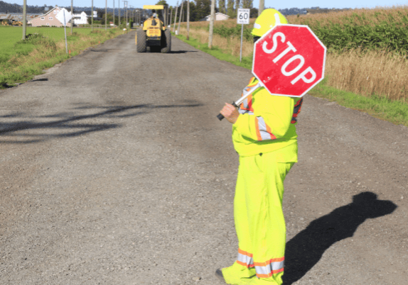 A construction worker holding a stop sign on a dirt road, with loader/tractor on the road up ahead.