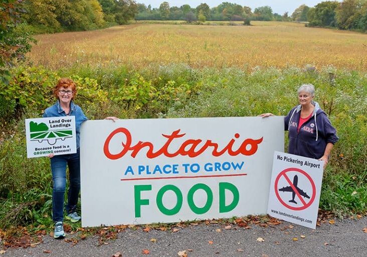 Two women standing next to a sign that says Ontario is a place to grow food on a farm.