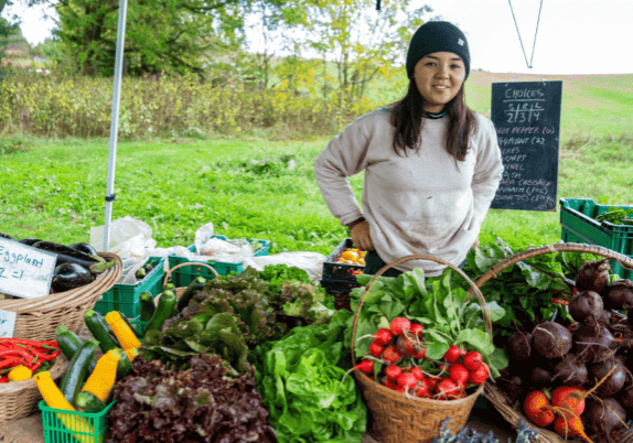 A woman under a tent standing behind a table with baskets of vegetables. Selling vegetables at a market.