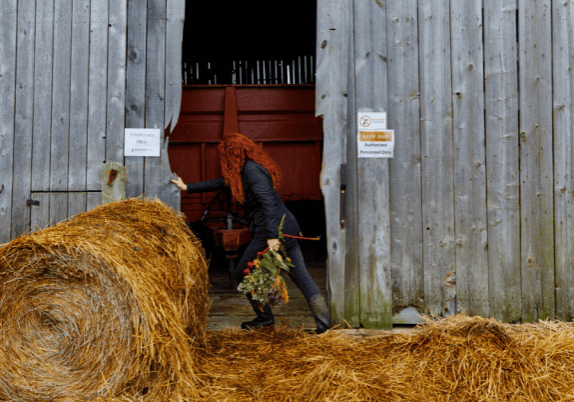 A woman carrying a bouquet of flowers opening doors to a barn with a bale of hay in front of the barn and hay on the ground.