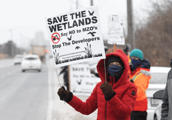 A group of people holding signs that say save the wetlands, say no to MZOs, and stop the developers.