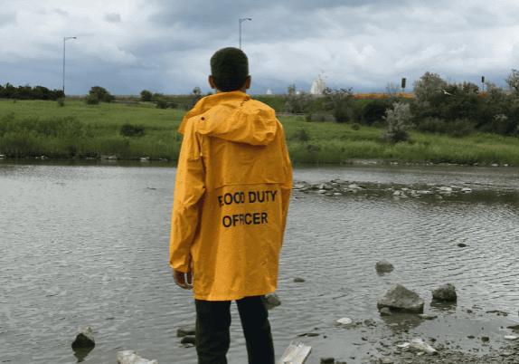 A person in a yellow raincoat that says "food duty officer" on the back, standing near a body of water.