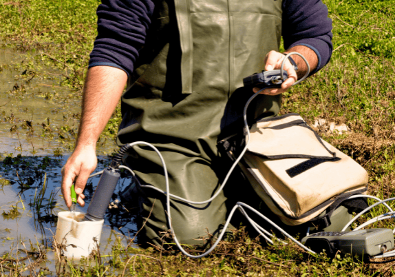 A man kneeling in a puddle testing groundwater with a small, portable machine.