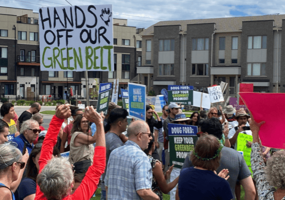 A group of people holding signs in front of a building "Hands off our Greenbelt".